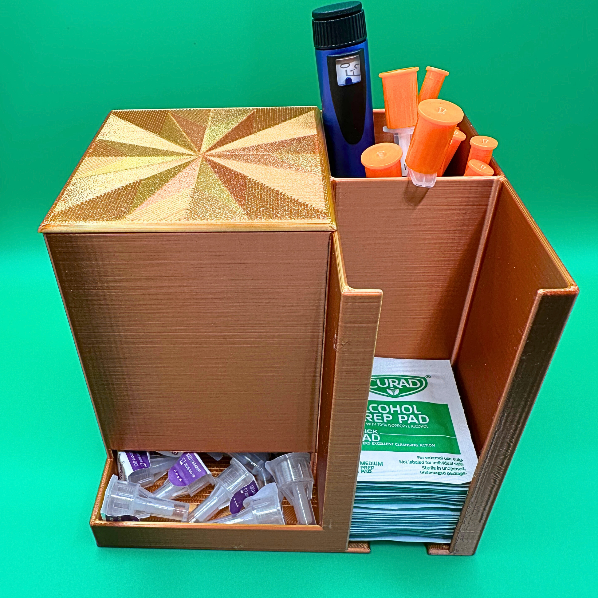 Bronze desktop injection organizer shown fully loaded with daily supplies, featuring a large left compartment holding a full box of injection pen needles with the lid in place, a rear right compartment holding an injection pen and capped syringes upright, and a front right compartment stacked with alcohol prep pads. Compact open-top organizer designed for desk or countertop use, keeping supplies visible, separated, and easy to access.