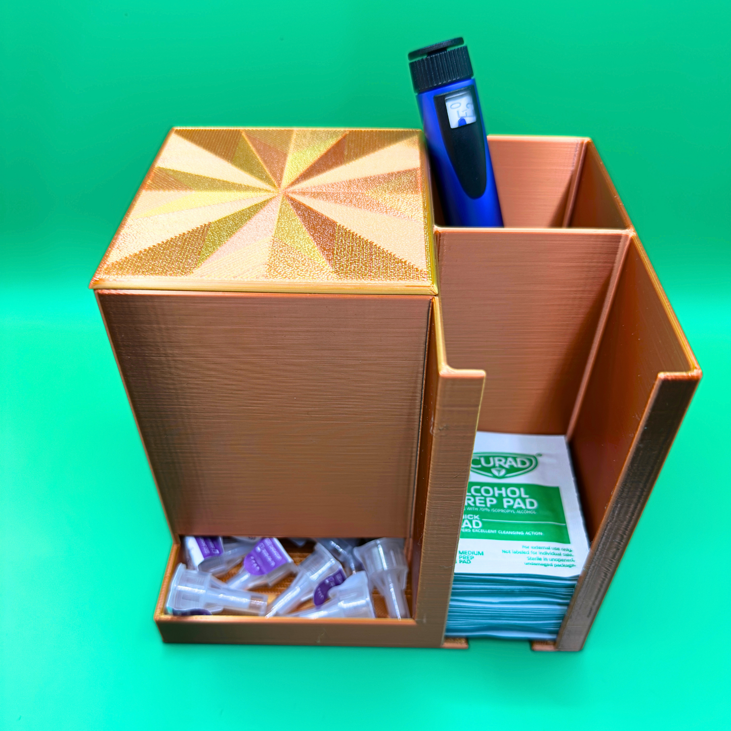 Bronze desktop injection organizer shown with the lid in place, featuring a large left compartment partially filled with injection pen needles, a rear right compartment holding an injection pen upright, and a front right compartment stacked with alcohol prep pads. Compact open-top organizer designed for desk or countertop use, keeping supplies organized, visible, and easy to access.