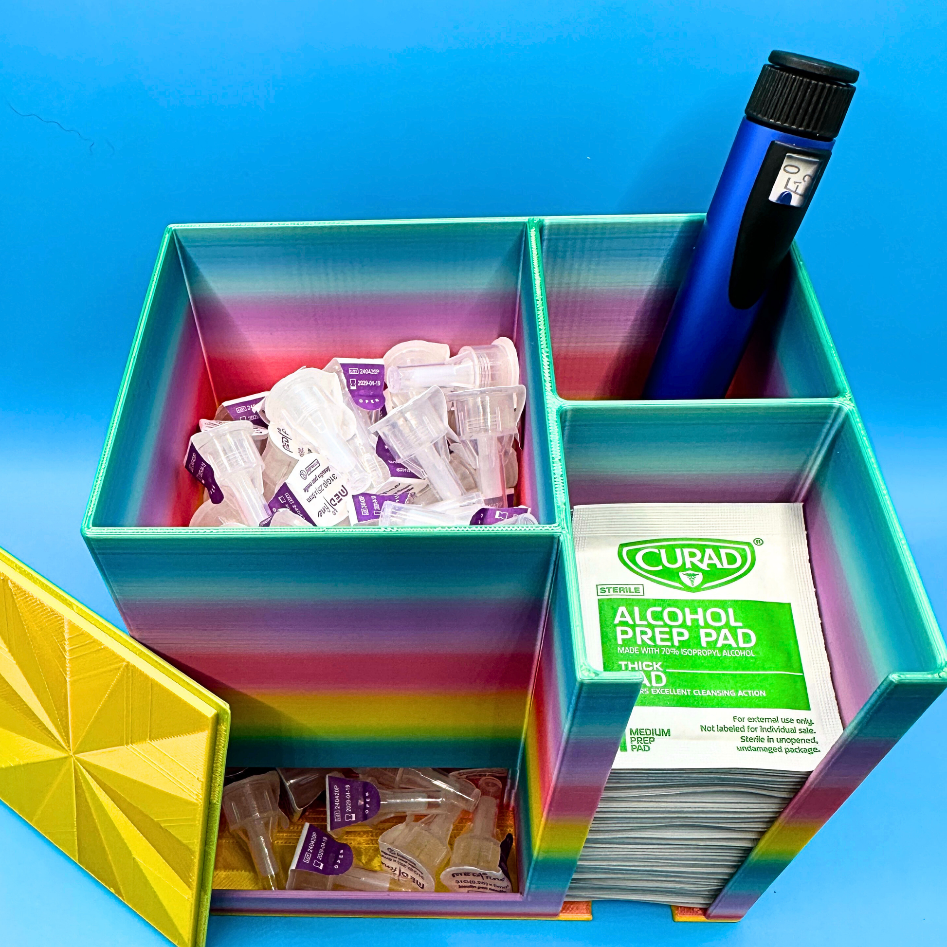 Open desktop injection organizer showing the internal layout, with a large left compartment filled with injection pen needles, a rear right compartment holding an injection pen upright, and a front right compartment stacked with alcohol prep pads. Compact open-top organizer designed for desk or countertop use, keeping needles, pens, and wipes organized, visible, and easy to access.