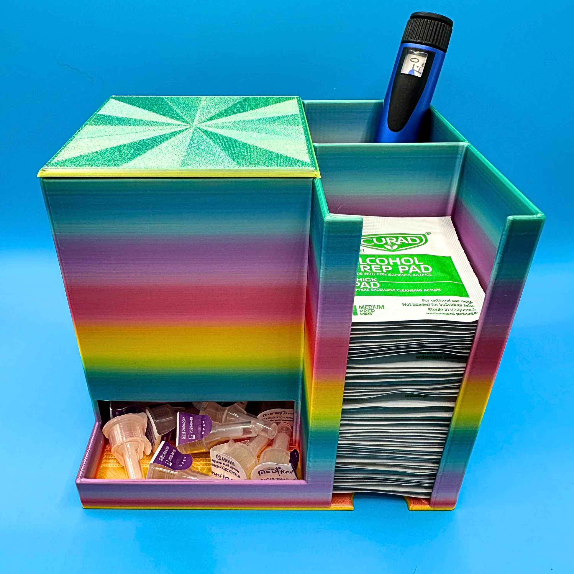 Desktop injection organizer with a large left compartment holding a full box of injection pen needles, a rear right compartment holding an injection pen upright, and a front right compartment stacked with alcohol prep pads. Compact open-top desktop organizer designed to keep needles, pens, and wipes organized and easy to access on a desk, countertop, or nightstand.
