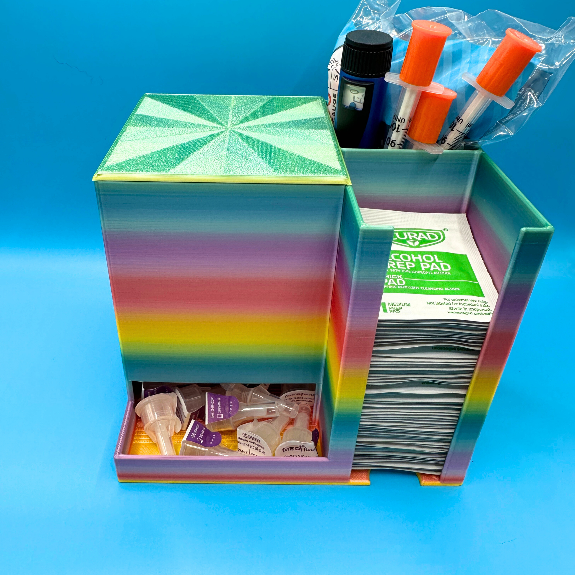 Rainbow desktop injection organizer shown fully loaded with supplies, featuring a large left compartment holding a full box of injection pen needles with the lid in place, a rear right compartment holding an injection pen and packaged syringes upright, and a front right compartment stacked with alcohol prep pads. Compact open-top organizer designed for desk or countertop use, keeping supplies visible, separated, and easy to access.